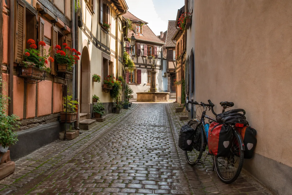 Ruelle pavée d’un village viticole alsacien avec vélo chargé de sacoches appuyé contre un mur